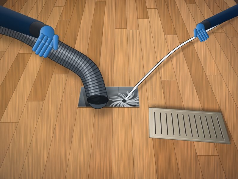 Gloved hands feeding a flexible vacuum hose and a spinning rotary brush into an open floor supply duct, with the register cover set aside on the hardwood floor