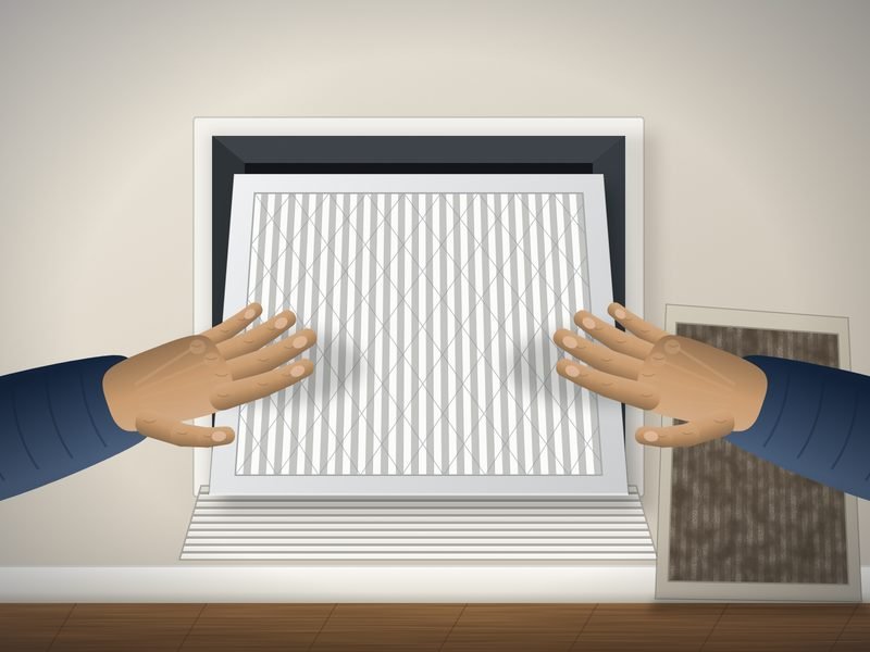 Hands sliding a clean pleated air filter into an open wall return grille, with a dirty used filter leaning against the wall below